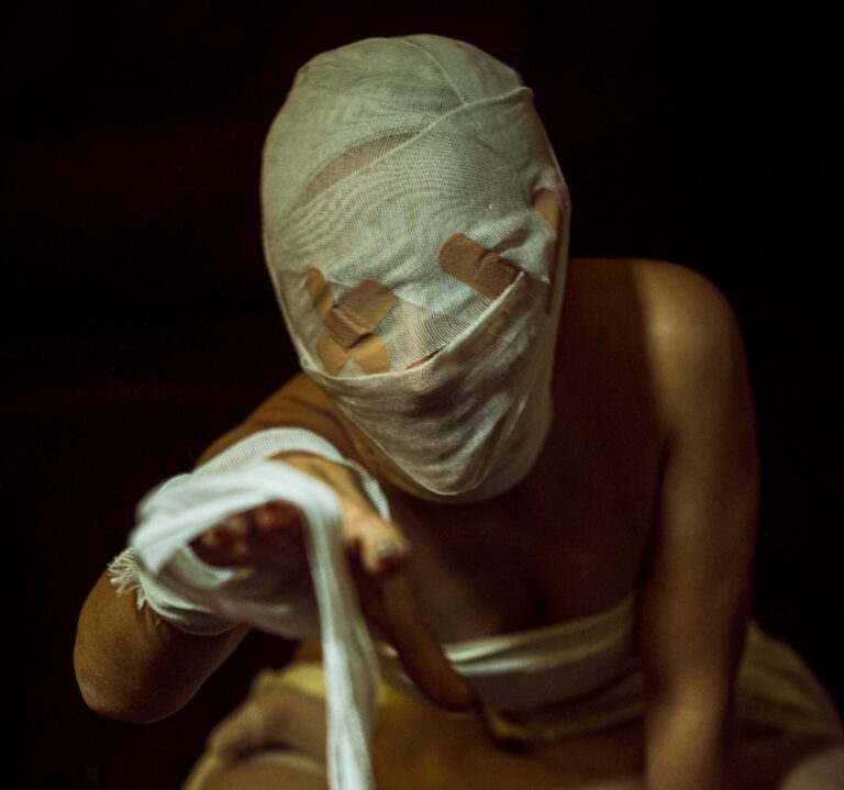 A person wrapped in bandages extends a hand in a dimly-lit setting, evoking mystery.