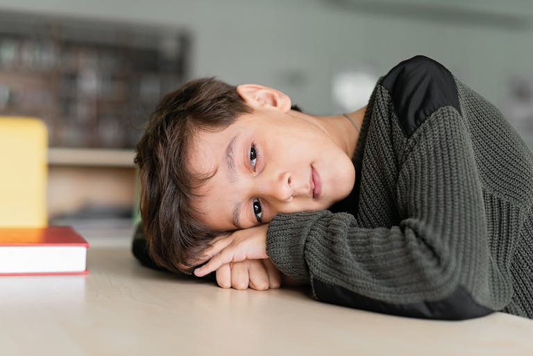 A young boy in a black sweater lies on a desk, looking pensively indoors.