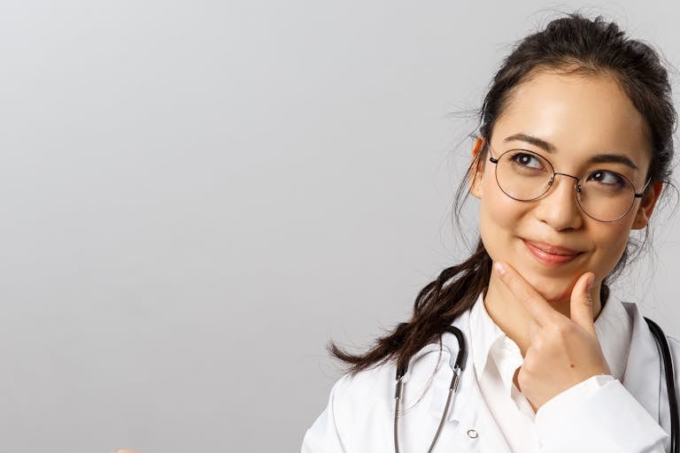 Asian female doctor wearing glasses, smiling thoughtfully with hand gesture on chin.