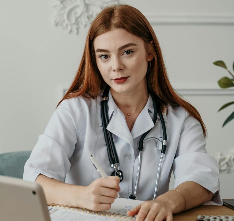 Portrait of a young female doctor with stethoscope, taking notes at her desk with an electronic tablet.